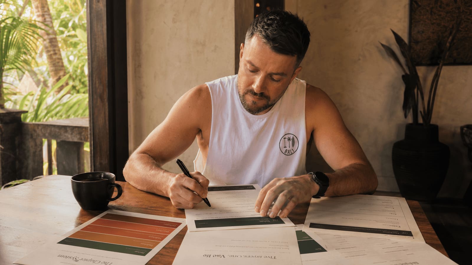 Jason filling in the printed Chapter System sheets at a wooden desk in Bali, morning light, coffee beside him.